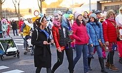 Eine Gruppe von Menschen in verschiedenen Kostümen geht auf einer Straße, einige tragen Babys und andere spielen Instrumente. Bäume und Gebäude sind im Hintergrund.