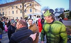 Eine Gruppe von Menschen in Kostümen geht auf einer Straße, einer in einem Hulk-Kostüm, und ein Mann mit Brille. Gebäude sind im Hintergrund sichtbar.