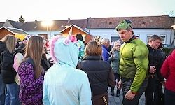 Eine Gruppe von Menschen ist in einem Außenbereich versammelt, einige in Kostümen. Ein Mann in einem grünen Hulk-Kostüm steht vor einer Frau in einem Einhorn-Kostüm. Sie stehen vor einem Gebäude mit mehreren Fenstern.