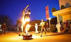 Eine Nachtszene mit einem großen Lagerfeuer und einer Person in Kostüm, die eine Fackel hält. Eine große Statue steht hinter dem Feuer, und andere in weißen Outfits sind in der Nähe. Das Gebiet ist durch Straßenlaternen und die untergehende Sonne beleuchtet.