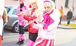 Zwei Frauen in rosa und weißen Kostümen spielen Flöten auf der Straße. Ein Auto und eine Person sind im Hintergrund.