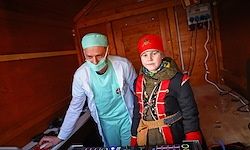 Eine Person in medizinischer Kleidung steht neben einer jungen Person in einem Piratenkostüm, beide stehen vor einer DJ-Anlage in einem Holzraum.