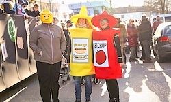 Drei Personen in Kostümen stehen auf einer Straße. Eine Person trägt ein Ketchupflaschen-Kostüm, eine andere ein Senfflaschen-Kostüm und die dritte ein Lego-Mann-Kostüm. Im Hintergrund sind Menschen und Autos zu sehen.
