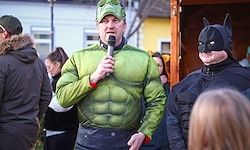 Ein Mann in einem grünen Hulk-Kostüm spricht in ein Mikrofon, umgeben von einer Menschenmenge, darunter ein Mann in einem Batman-Kostüm.