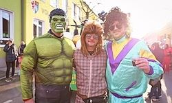 Drei Menschen in Kostümen posieren für ein Foto auf einer Stadtstraße mit Gebäuden im Hintergrund. Eine Person trägt ein Hulk-Kostüm, eine andere eine rosa Perücke und die dritte ein blaues und gelbes Kostüm.