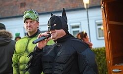 Ein Mann in einem Batman-Kostüm singt in ein Mikrofon, während ein anderer Mann in einem grünen Kostüm hinter ihm steht. Sie stehen vor einem Gebäude.