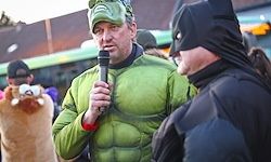 Ein Mann in einem Hulk-Kostüm spricht in ein Mikrofon, während ein anderer Mann in einem Batman-Kostüm zuhört. Sie befinden sich in einem öffentlichen Bereich mit einem Bus im Hintergrund.