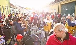 Eine überfüllte Straße mit Menschen in Kostümen und Hüten. Eine Person in einer roten Jacke geht mit einem Kinderwagen. Die Sonne scheint hell und Gebäude säumen die Straße.