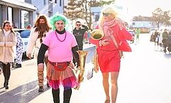 Menschen in Kostümen gehen auf der Straße. Ein Mann spielt Posaune, und ein anderer trägt ein rosa Shirt. Im Hintergrund sind Gebäude und Bäume zu sehen.