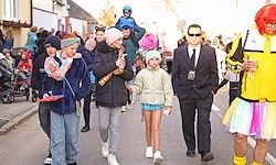 Eine Gruppe von Menschen geht die Straße entlang. Dazu gehören ein Mann in Anzug, ein Kind mit rosa Hut, eine Frau mit Baby und eine Person in einem leuchtend gelben Kostüm.