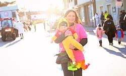 Eine Frau in einem rosa Jacke hält ein Kind in einem gelben Kostüm mit einem Schwanz. Sie stehen auf einer Straße mit Fußgängern, Gebäuden und einem Fahrzeug im Hintergrund.