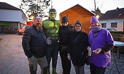 Fünf Personen stehen in einem Wohngebiet, drei sind als Superhelden verkleidet. Der Hulk, Batman und eine in Lila gekleidete Person. Hinter ihnen sind Häuser, ein Tisch und ein geparktes Auto.