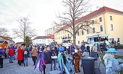 Eine Menschenmenge, einige in Kostümen, steht vor einem Gebäude mit roten Dächern. Tische, Mülleimer und ein Bus sind vorhanden. Bäume und ein bewölkter Himmel sind sichtbar.