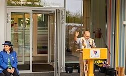 Ein Mann in einem bunten Outfit spricht an einem Podium vor einem Gebäude. Eine Frau in einer blauen Jacke sitzt in der Nähe. Das Gebäude hat Glastüren und -wände.
