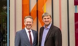 Zwei Männer in Anzügen und Krawatten stehen nebeneinander vor einem Gebäude mit mehrfarbigen Wandpaneelen.