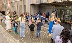 Eine Gruppe von Menschen, einschließlich Kinder, stehen und sitzen vor einem Gebäude. Einige Erwachsene interagieren mit den Kindern, möglicherweise bei einer Aktivität. Die Umgebung scheint ein Outdoor-Event mit Ziegelboden und einem Garten zu sein.