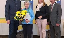 Eine Gruppe von vier Erwachsenen steht vor einem Hintergrund, die Frau in der Mitte hält einen Strauß gelber Blumen und ein Dokument. Die anderen drei sind in formeller Kleidung gekleidet.
