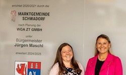 Zwei Frauen posieren vor einer Wand mit dem Schild 'Marktgemeinde Schwadorf'. Sie lächeln, eine trägt eine Halskette. Ein Wappen ist auf der linken Seite.