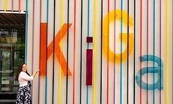 Eine Frau steht neben einer farbenfrohen Wand mit großen Buchstaben, die 'KIGa' bilden. Sie zeigt mit einem leuchtenden und fröhlichen Ausdruck auf die Buchstaben.