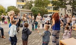 Eine Gruppe von Erwachsenen und Kindern versammelt sich vor einem Gebäude. Einige tanzen, während andere zuschauen. Die Kinder tragen verschiedene Kleidungsstücke und Accessoires. Bäume und ein Van sind im Hintergrund.
