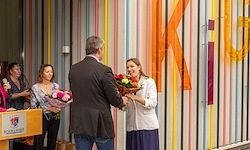 Eine Frau erhält Blumen von einem Mann, der vor einer mehrfarbigen Wand mit großen Buchstaben steht. Eine Gruppe von Menschen beobachtet sie.