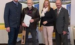 Vier Menschen stehen eng beieinander und halten ein Zertifikat, ein Papier und einen Beutel. Sie stehen vor einem weißen und gelben Hintergrund. Sie sind alle formell gekleidet.