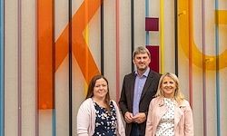 Drei Personen stehen vor einer Wand mit einem großen Buchstaben K und bunten Streifen. Die beiden Frauen tragen helle Jacken, während der Mann einen dunklen Anzug trägt.