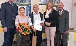Eine Gruppe von Menschen steht zusammen und posiert für ein Foto. Die Frau in der Mitte hält einen Blumenstrauß und ein Zertifikat. Der Mann rechts trägt einen Anzug. Der Mann links trägt einen blauen Mantel.