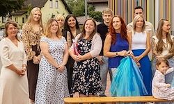 Eine Gruppe von Menschen steht vor einem Gebäude und posiert für ein Foto. Frauen in verschiedenen Kleidern, eine hält ein Kind, und ein Mann in einem schwarzen T-Shirt. Bäume und ein Haus im Hintergrund.
