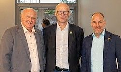Drei formell gekleidete Männer stehen zusammen, wahrscheinlich für ein Foto. Der Mann links trägt einen grauen Anzug, der in der Mitte einen schwarzen Anzug und der rechts einen marineblauen Anzug.