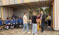 Zwei Männer stehen vor einem Podium und tauschen etwas aus. Dahinter sitzt eine Gruppe von Menschen auf einer Bank, einige stehen in der Nähe. Die Atmosphäre ist formell, mit einem Gebäude im Hintergrund.