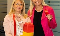 Zwei Frauen lächeln und posieren für ein Foto, eine hält ein rotes Band und die andere einen großen Lego-Baustein. Sie sind in Pink bzw. Schwarz gekleidet, der Hintergrund scheint ein Büro zu sein.