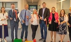 Eine Gruppe von Menschen, feierlich gekleidet, steht in einem Raum und hält Bänder, wahrscheinlich zur Eröffnung. Ein Lego-Karton liegt auf dem Boden.
