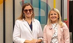 Zwei Frauen stehen nebeneinander, lächeln und posieren für ein Foto vor einer bunten Wand mit einem großen Buchstaben 'D'.