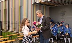 Ein Mann im Anzug reicht einer Frau in einem weißen Oberteil einen Blumenstrauß vor einer Gruppe von Menschen in blauen Uniformen.