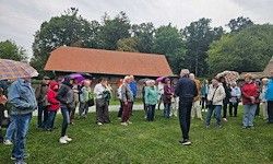 Eine Gruppe von Menschen steht auf einem Rasenfeld und hält Regenschirme, mit einem traditionellen Haus mit rotem Dach im Hintergrund, unter bewölktem Himmel.