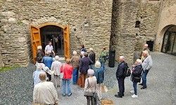 Eine Gruppe von Menschen steht vor einem historischen Steingebäude mit einer offenen Holztür, wahrscheinlich Touristen. Sie sind lässig gekleidet und einige tragen Taschen. Die Umgebung ist sonnig.