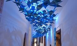 Ein Flur mit weißen Wänden ist mit einer blauen Origami-Vogel-Deckenleuchte dekoriert. Der Raum ist schwach beleuchtet.