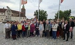 Eine Gruppe älterer Menschen posiert vor einem Schloss mit Fahnen und einer Statue. Sie sind in verschiedenen Farben gekleidet und einige tragen Handtaschen.