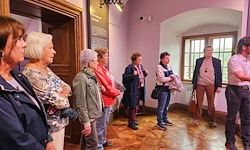 Eine Gruppe von Menschen steht in einem Raum mit Holzböden, rosa Wänden und großen Fenstern. Einige tragen Jacken und Schuhe, und zwei unterhalten sich.