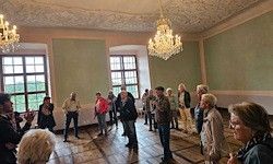 Eine Gruppe von Menschen steht in einem Raum mit Kronleuchtern, Holzböden und großen Fenstern, möglicherweise an einer Besprechung oder einer Tour teilnehmend.
