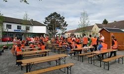 Eine große Gruppe von Menschen in orangen Westen sitzt an Tischen vor einem Gebäude mit Bäumen und Häusern im Hintergrund.