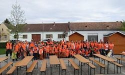 Eine Gruppe von Menschen in orangen Westen und Helmen versammelt sich vor einem Gebäude mit mehreren Fenstern und Türen und posiert für ein Foto.