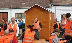 Eine Gruppe von Menschen in orangefarbenen Westen steht draußen, einige in der Nähe eines Holzschuppens und andere auf Bänken sitzend, im Gespräch.
