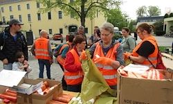 Freiwillige in orange Westen bereiten Essen zur Verteilung vor. Eine Gruppe von Menschen steht um einen Tisch mit Essensboxen und Tüten. Ein Kind sitzt auf einer Bank in der Nähe eines Gebäudes.