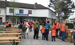 Eine Gruppe von Menschen in orangenen Westen steht vor einem Gebäude mit dem Namen 'Sparkasse'. Einige schauen auf einen Tisch mit Kisten, während andere sprechen. Bäume und ein klarer Himmel sind im Hintergrund.