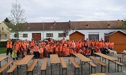 Eine große Gruppe von Menschen in orangefarbenen Westen versammelt sich um Holzbänke vor einem Gebäude.