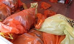 Mehrere orange und gelbe Plastiktüten, gefüllt mit Müll, sind auf einem roten Boden verstreut. Die Tüten scheinen von einer Müllsammlung zu stammen.