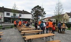 Eine Gruppe von Menschen in orangenen Westen versammelt sich um Holztische auf einer Kopfsteinpflasterstraße, mit mehreren Gebäuden und Bäumen im Hintergrund.