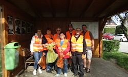 Eine Gruppe von Personen in Warnwesten steht in einem Schuppen und hält Taschen. Ein Mülleimer befindet sich in der Nähe, und ein Auto ist draußen geparkt.